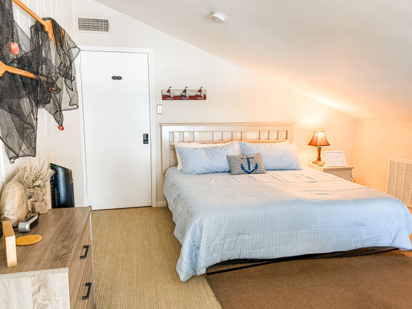 Cozy sunlit coastal attic bedroom with sloped ceiling, king bed dressed in blue-and-white striped bedding, anchor throw pillow, wooden headboard, bedside lamp and nautical accents.