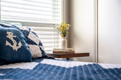 Sunlit nautical-themed bedroom corner with navy anchor pillow and blue-and-white quilt, wooden nightstand with stacked books and a vase of yellow flowers beside white window blinds.