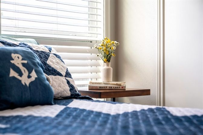 Sunlit nautical-themed bedroom corner with navy anchor pillow and blue-and-white quilt, wooden nightstand with stacked books and a vase of yellow flowers beside white window blinds.
