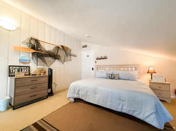 Cozy lake house attic bedroom with sloped ceiling, king bed in blue striped linens, nautical paddle and fishing-net wall decor, wooden dresser and bedside lamp.