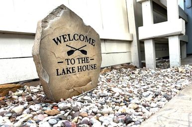 Weathered decorative stone marker engraved "WELCOME - TO THE - LAKE HOUSE" with crossed oars, nestled among multicolored pebbles beside white house siding and porch steps.
