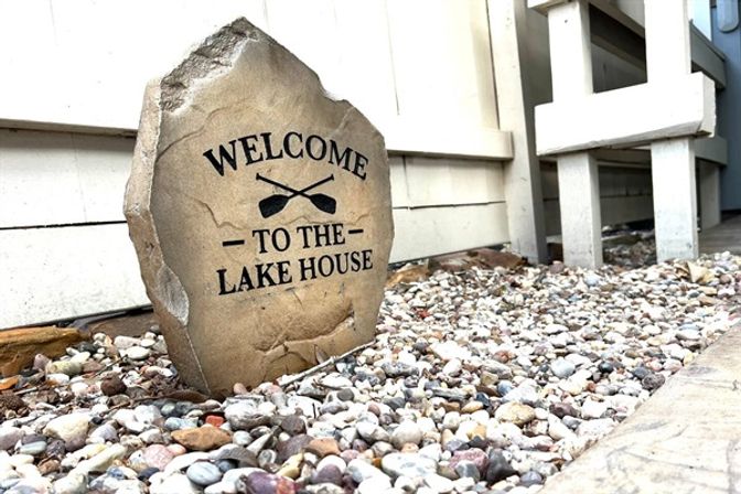 Weathered decorative stone marker engraved "WELCOME - TO THE - LAKE HOUSE" with crossed oars, nestled among multicolored pebbles beside white house siding and porch steps.