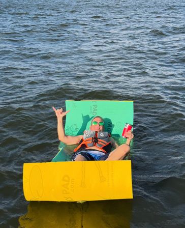 Person in a life jacket and sunglasses lounging on bright green and yellow foam pads, floating on a lake, holding a red can and flashing a shaka sign — relaxed summer water scene.
