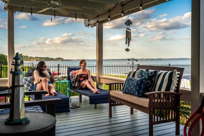 Lakefront covered porch with two people lounging on navy cushioned chairs, a wooden bench with nautical pillows, string lights and wind chimes overhead, and a wide waterfront view of a calm lake and distant shoreline.