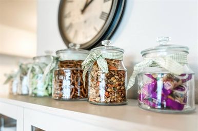Decorative glass jars tied with green gingham ribbons, filled with pretzels, mixed nuts and wrapped candies, lined up on a white sideboard beneath a large wall clock.