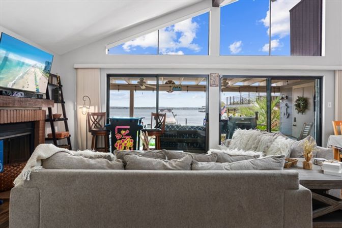 Bright waterfront living room with gray sectional, vaulted windows and sliding glass doors opening to a covered patio and dock — relaxed coastal vibe.