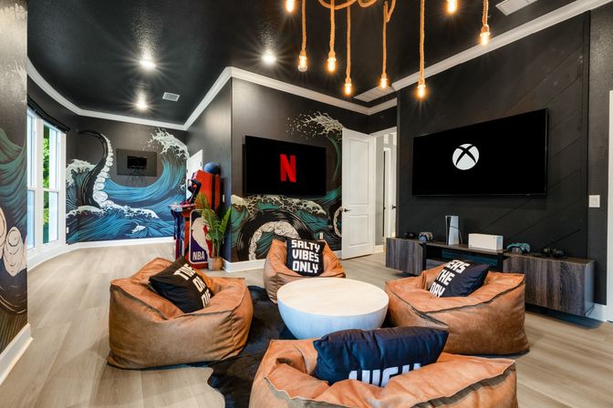 Modern coastal-themed gaming and media room with octopus wave wall mural, hanging Edison bulbs, two wall TVs showing Netflix and Xbox logos, leather beanbag chairs around a round coffee table and an arcade cabinet.