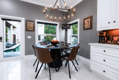 Modern kitchen dining nook with round black table set for four, brown leather chairs, industrial chandelier, bowl of oranges, white cabinetry and backyard pool view.