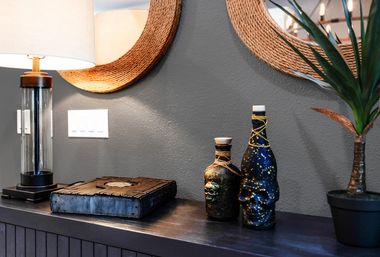 Modern entryway vignette on a dark console: lit glass table lamp, woven round mirror on a gray textured wall, rustic book, quirky skull-accented and star-speckled decorative bottles, and a potted faux plant.