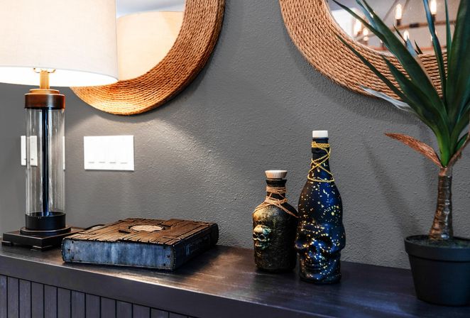 Modern entryway vignette on a dark console: lit glass table lamp, woven round mirror on a gray textured wall, rustic book, quirky skull-accented and star-speckled decorative bottles, and a potted faux plant.