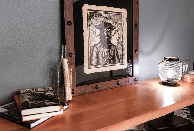 Cozy wooden desk vignette with a framed vintage pirate engraving, glass bottle with a rolled note, stacked books and a glowing mesh lantern against a gray wall.