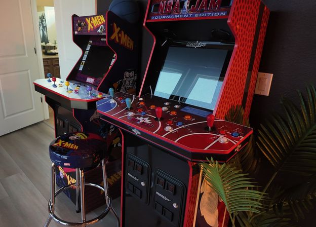 Vibrant home game room corner with two retro arcade cabinets (X-Men and NBA Jam), red-and-black control panels with colorful joysticks and a matching X-Men stool on light wood floor.