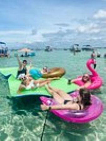 Group of people lounging on colorful inflatables — pink flamingo, donut and green mat — floating in clear shallow turquoise water with boats in the distance under a blue sky.