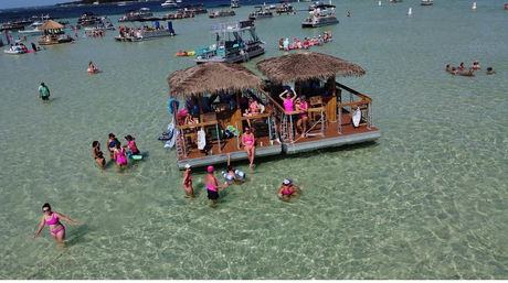 Aerial view of a floating tiki-pontoon bar at a lively sandbar party, people in bright swimsuits wading in crystal-clear shallow turquoise water with boats nearby.