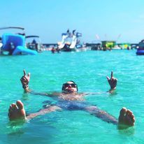 Person floating on back in clear turquoise water near beach boats and inflatables, wearing sunglasses and playfully showing both middle fingers.