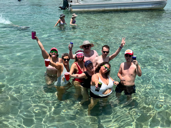 Group of friends partying waist-deep in clear turquoise water near an anchored boat, wearing swimsuits and sunglasses and holding drinks on a sunny beach day