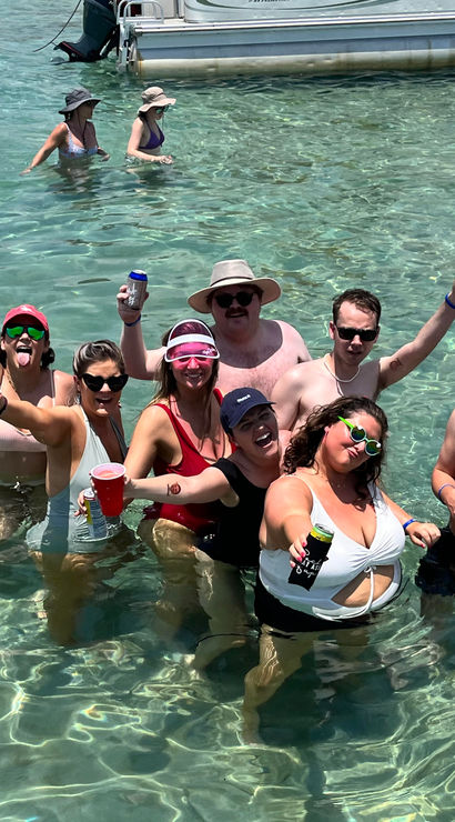 Group of friends partying waist-deep in clear turquoise water near an anchored boat, wearing swimsuits and sunglasses and holding drinks on a sunny beach day