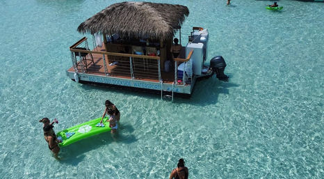 Aerial view of a floating tiki-bar pontoon in shallow crystal-clear turquoise water with people in swimsuits wading and playing on a bright green inflatable raft — tropical beach vibes.