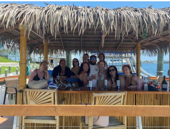 Smiling group posing at a bamboo tiki-style dock bar with a thatched roof, drinks on the counter and marina boats in the sunny waterfront background.