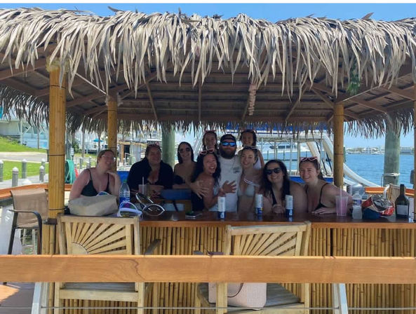 Smiling group posing at a bamboo tiki-style dock bar with a thatched roof, drinks on the counter and marina boats in the sunny waterfront background.