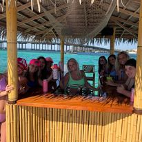 Cheerful group of friends at a round bamboo tiki bar with a thatched roof set over a turquoise tropical lagoon, with overwater bungalows and a wooden pier in the background.