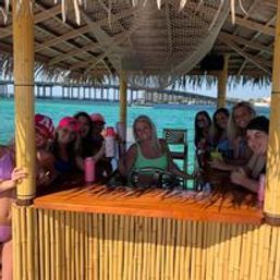 Cheerful group of friends at a round bamboo tiki bar with a thatched roof set over a turquoise tropical lagoon, with overwater bungalows and a wooden pier in the background.
