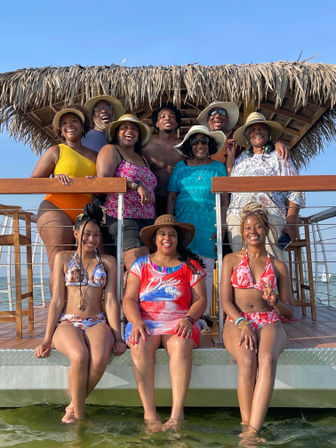 Smiling multigenerational group in swimsuits and sun hats posing under a thatched tiki hut on a sunny coastal pier with feet dipped in the water, beach vacation vibes.
