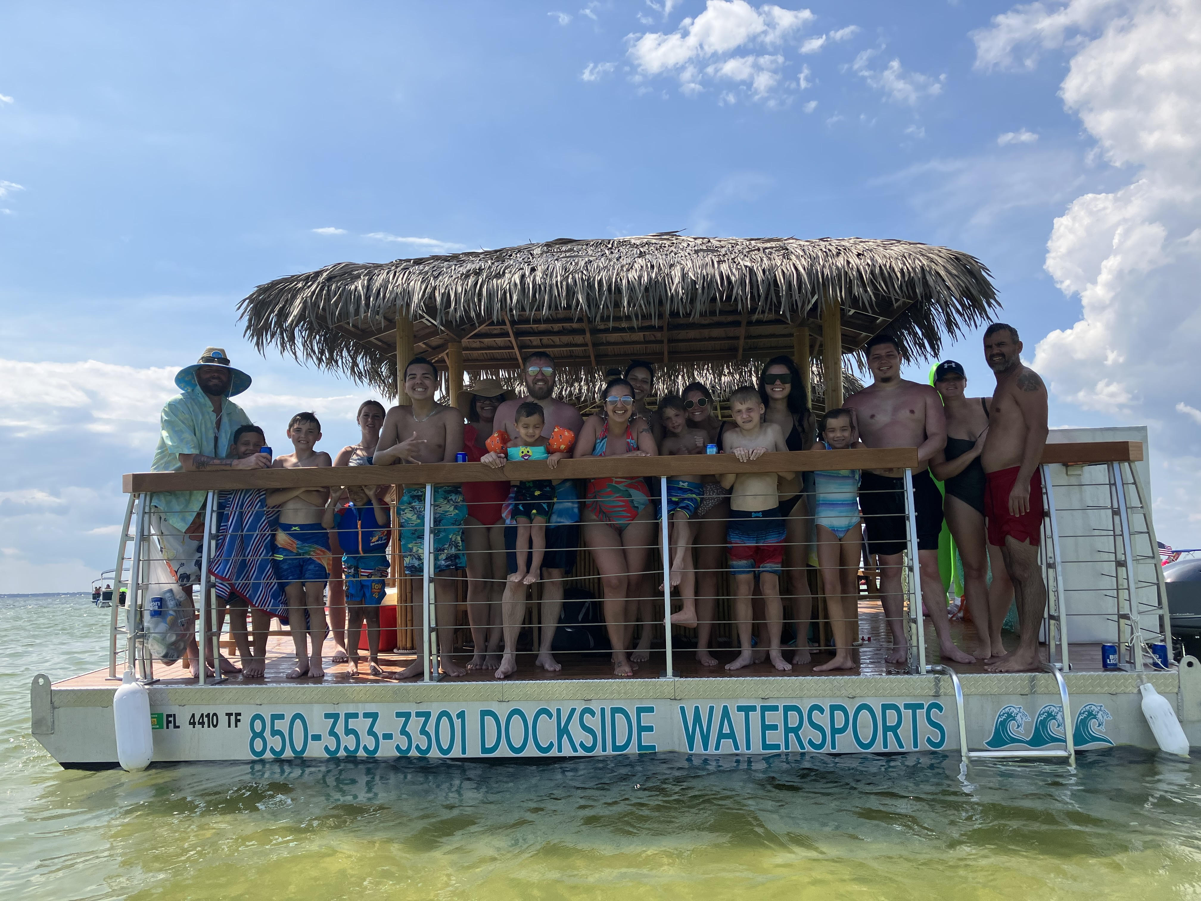 Large multigenerational group in swimsuits smiling under a thatched-roof tiki on a pontoon over clear shallow Gulf waters off the Florida Panhandle coast