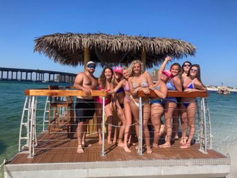 Group of friends in swimsuits posing on a thatched-roof tiki raft over turquoise bay with a coastal bridge and clear blue sky