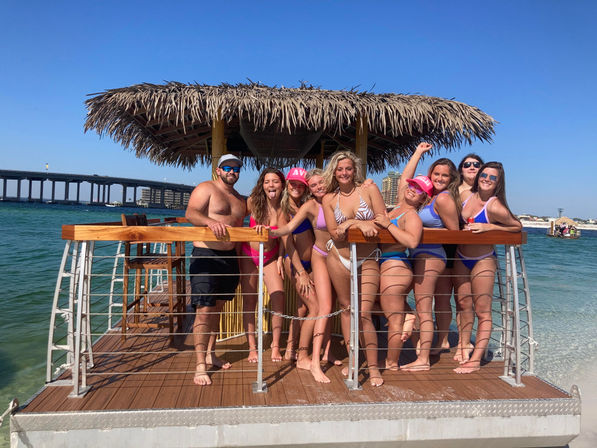 Group of friends in swimsuits posing on a thatched-roof tiki raft over turquoise bay with a coastal bridge and clear blue sky