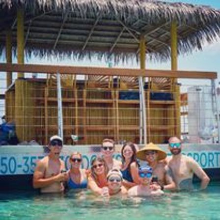 Smiling group of friends in clear turquoise water gathered around a thatched‑roof floating bar on a tropical boat tour, wearing sunglasses, sun hats and swimwear.