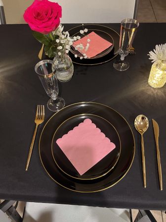 Elegant black-and-gold tablescape with stacked black plates, scalloped pink napkin, gold cutlery and champagne flutes, bright pink rose and baby’s breath in a glass vase, and a mason-jar fairy-light centerpiece.
