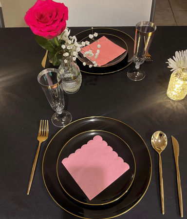 Romantic black-and-gold tablescape with scalloped pink napkin on black plates, gold flatware, champagne flutes, pink rose and baby's-breath centerpiece and a glowing jar light