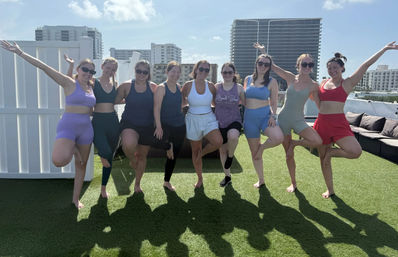 Ten women in colorful activewear strike tree pose on a sunny rooftop turf with coastal high-rise skyline in the background, a fun group fitness photo.