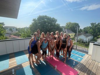 Sunny rooftop yoga session on a wooden deck in a leafy suburban neighborhood — a smiling group of women posing on colorful yoga mats with water bottles, trees and houses in the background.