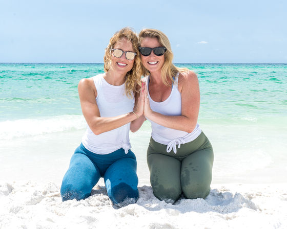 Two women in white tops and colorful leggings kneeling in a beach-yoga pose on white sand, palms together and smiling with turquoise ocean and clear sky behind them.