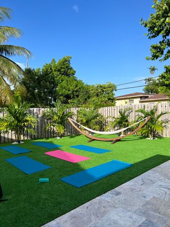 Sunny tropical backyard with artificial turf set up for yoga — blue and one pink mats, wooden hammock stand, palm plants and a wooden fence under a clear blue sky