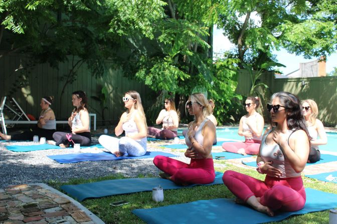 Sun-dappled outdoor yoga and breathwork session by a backyard pool — people in colorful leggings sit cross-legged on mats with hands on their chest, meditating under leafy trees.