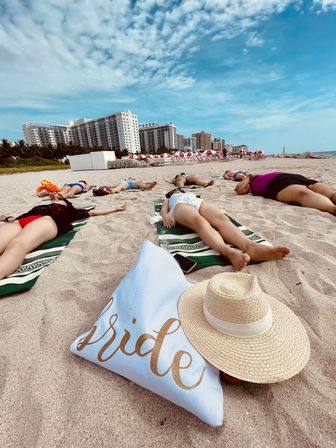 Bachelorette beach scene at a coastal resort: friends sunbathing on towels with a glitter 'bride' pillow and straw hat in the foreground, high-rise hotels and blue sky along the shoreline