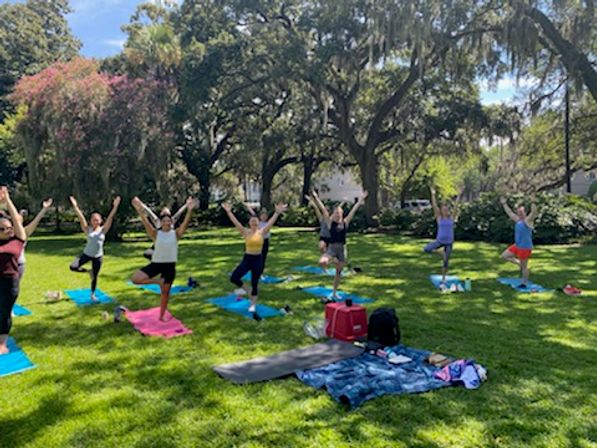 Outdoor yoga class with a group holding tree pose on colorful mats in a sunny park beneath large oak trees, water bottles and blankets on the grassy lawn