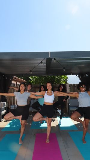 Outdoor backyard yoga class on a sunny patio under a shaded pergola — six women balancing in tree pose on colorful yoga mats, smiling with arms outstretched.