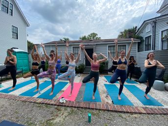 Outdoor backyard yoga class with a group balancing in tree pose on colorful mats in a residential yard under a partly cloudy sky.