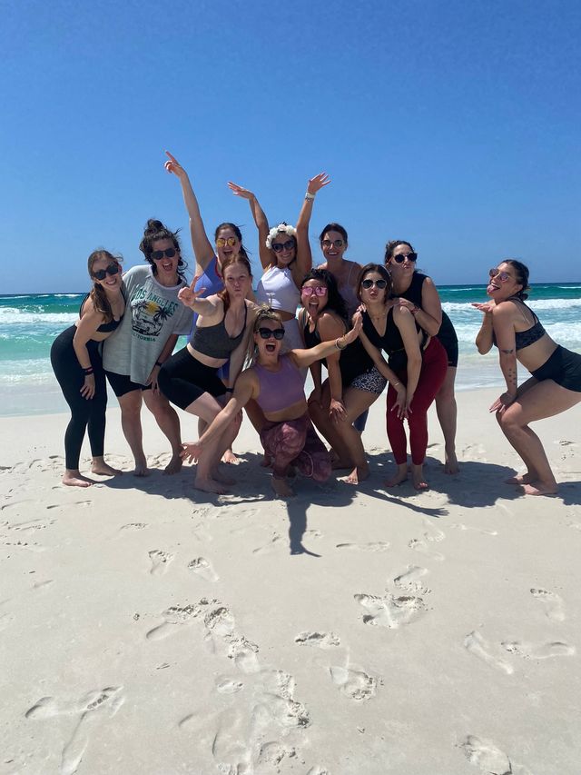 Group of friends in activewear and swimsuits laughing and posing on a white-sand beach with turquoise ocean waves and a clear blue sky.