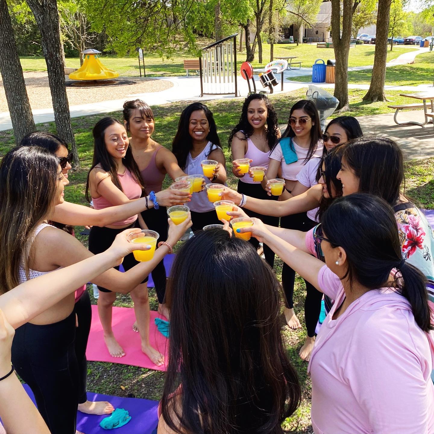 Group of friends standing in a circle on colorful yoga mats at a sunny park picnic, toasting with cups of orange juice.