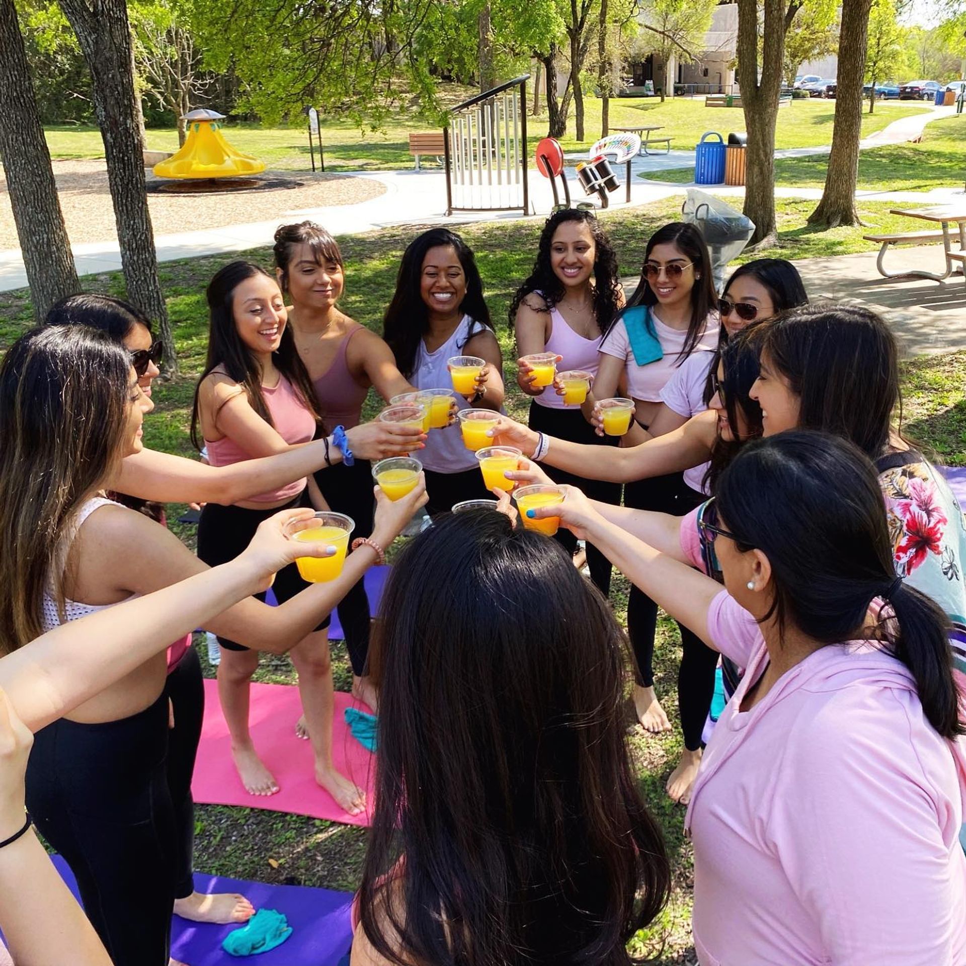 Group of friends standing in a circle on colorful yoga mats at a sunny park picnic, toasting with cups of orange juice.