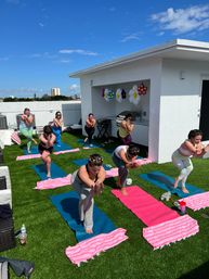 Outdoor rooftop yoga class on a sunny urban roof — participants balancing on colorful mats and pink striped towels on green turf under a bright blue sky with festive flower balloons.