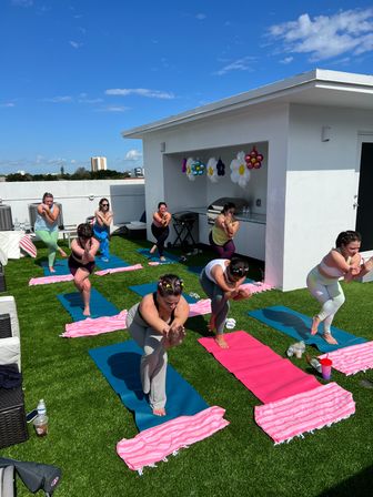 Outdoor rooftop yoga class on a sunny urban roof — participants balancing on colorful mats and pink striped towels on green turf under a bright blue sky with festive flower balloons.
