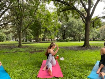 Outdoor yoga in a leafy park — woman doing a seated spinal twist on a pink mat on green grass, water bottle and shoes beside her with other mats and large trees in the background.