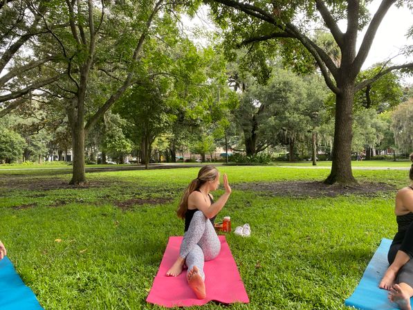 Outdoor yoga in a leafy park — woman doing a seated spinal twist on a pink mat on green grass, water bottle and shoes beside her with other mats and large trees in the background.