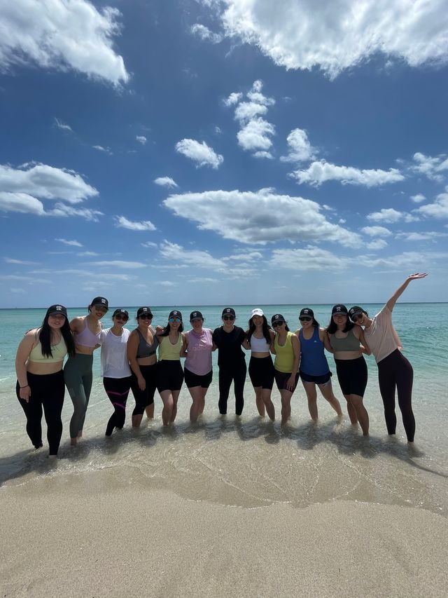 Thirteen friends standing waist-deep in clear turquoise ocean at a sunny beach, wearing activewear and caps under a bright blue sky dotted with fluffy clouds.